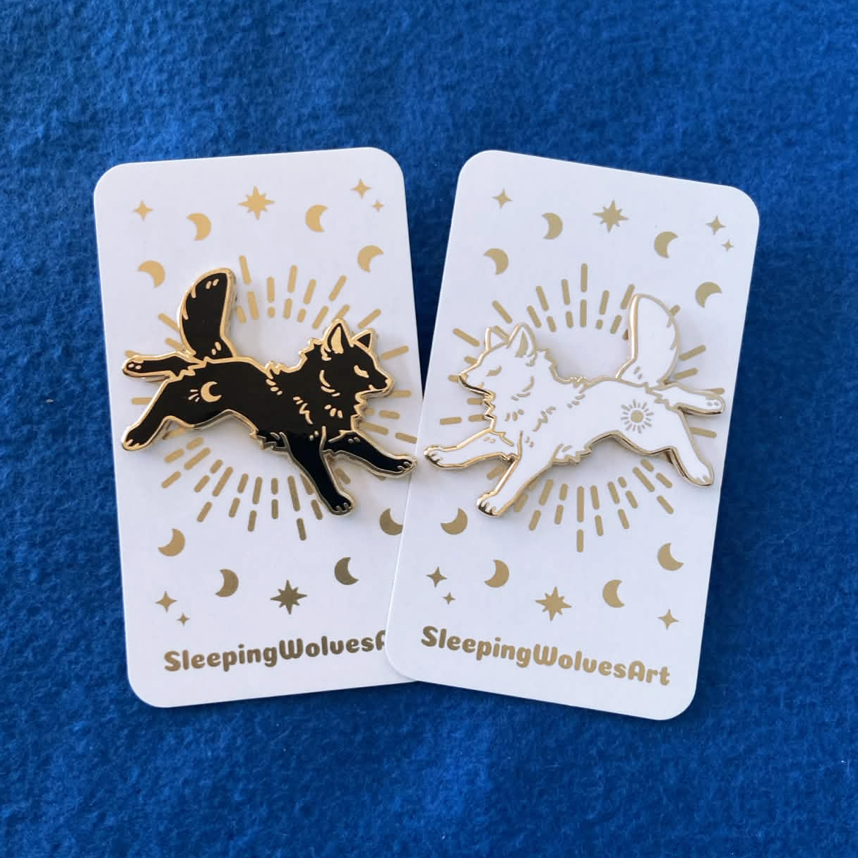 Two enamel pins of wolves on cards with 'SleepingWolvesArt' branding against a blue background.